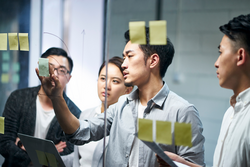 young asian entrepreneur of small company putting a adhesive note on glass in office during team meeting formulating business strategies.